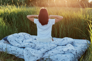 Slim female with dark hair wearing white casual t shirt posing backwards with raised arms, stretching hands after sleeping, enjoying sunrise in green meadow in beautiful nature.